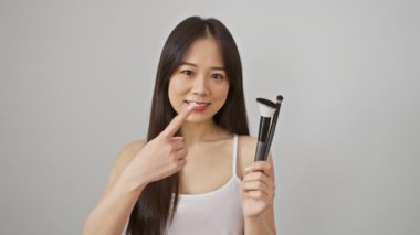 Neşeli genç Çinli kadın, en sevdiği makyaj fırçasını sunuyor, eli güvenle işaret ediyor, ağzı açık gülüşü, izole edilmiş beyaz arka planı güzellik ve pozitiflikle dolduruyor.
