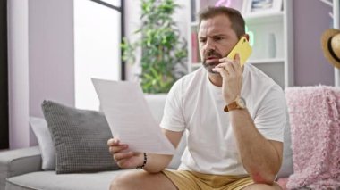 Orta yaşlı, gri saçlı bir adam, elinde modern bir oturma odasında elinde bir belge tutarken sarı bir telefonla konuşuyor..
