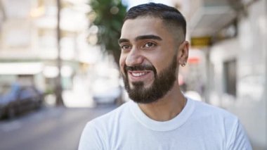 Güneşli bir caddede gülümseyen sakallı bir gencin portresi.