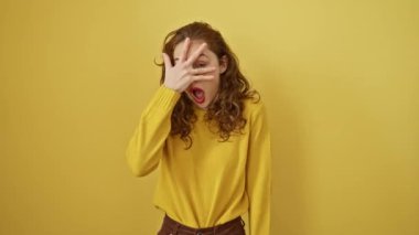Şok olmuş, genç, güzel bir kadın utangaçlıkla yüzünü gizliyor, parmaklarının arasından bakıyor, korkuyor ama vay canına, sarı arka planda izole edilmiş.