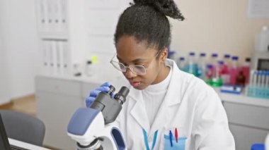 Odaklanmış bir siyah kadın bilim adamı modern bir laboratuvar ortamında mikroskop kullanarak örnekleri inceliyor..