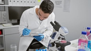 Laboratuvar önlüğü giymiş odaklanmış bir adam laboratuvar ortamında mikroskop altında bir kaydırağı inceliyor ve notlar yazıyor..