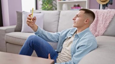 Rahat genç bir adam, rahat bir oturma odasındaki kanepede uzanırken akıllı bir telefonla selfie çekiyor..