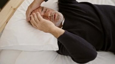 Olgun bir adam yatak odası ortamında kederli bir ifadeyle yatakta yatıyor, başını tutuyor..