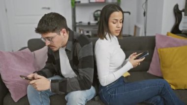 Bir adam ve bir kadın akıllı telefonlara dalmış, modern oturma odasındaki renkli minderlerle bir koltukta oturuyorlar..