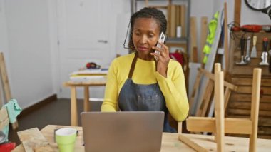 Afrikalı bir kadın ahşap atölyesinde dizüstü bilgisayar ve telefon kullanıyor.