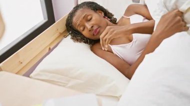 Olgun siyah bir kadın uyanıyor ve güneşli bir yatak odasında telefonuna uzanıyor.