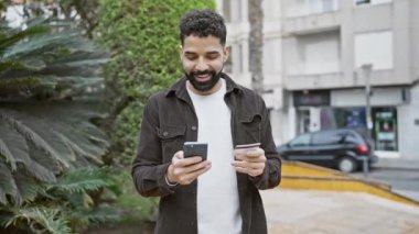 Şehir caddesinde elinde kredi kartı ve akıllı telefon tutan yakışıklı sakallı adam..