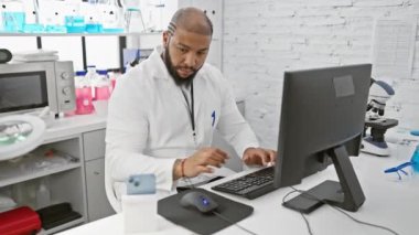 Laboratuvarda odaklanmış siyah bir adam akıllı telefon kullanırken bilgisayardaki verileri analiz ediyor.