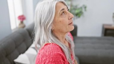 Güzel, orta yaşlı bir kadın, ciddi bir ifadeyle masum bir şekilde oturma odasındaki kanepesine yerleşir, rahatça dinlenir, evdeki yumuşak güneş ışığının altında güneşlenir..