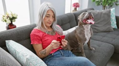 Neşeli, kır saçlı, orta yaşlı bir kadın evinde koltukta oturuyor, telefonuyla mesajlaşırken gülümsüyor, mutlu köpeğiyle eğleniyor.