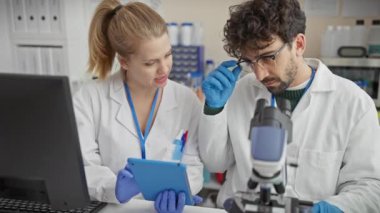 Laboratuvar önlüklü bir adam ve kadın bilimsel bir laboratuvar ortamında örnekleri mikroskop ve tabletle analiz ediyorlar..