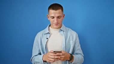 Mavi arka planda cep telefonu tutarken gülümseyen ve onay veren İspanyol asıllı genç adam..