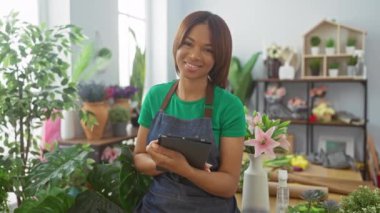 Önlüklü gülümseyen kadın canlı çiçekçide tablet tutuyor.