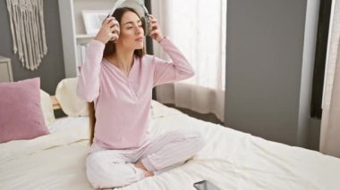 Pijamalı bir kadın kulaklıklı müzikten zevk alıyor ve rahat bir yatak odasında akıllı telefon kullanıyor..