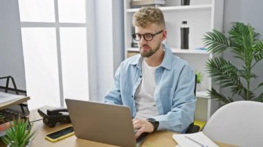 Modern ofis ortamında akıllı telefon ve laptop kullanan sakallı ve mavi gözlü yakışıklı bir genç..