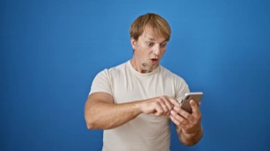 Sakallı, şaşırmış, beyaz bir adam mavi bir duvara karşı telefonunun içeriğine tepki veriyor..