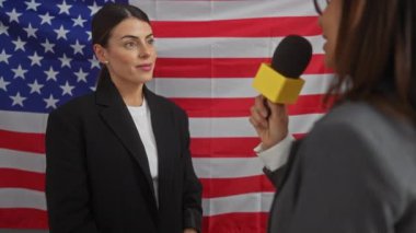 Bir kadın politikacı, bir kadın gazeteci tarafından bir ofisin içinde Amerikan bayrağı önünde sorgulanıyor..