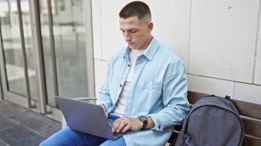 Modern bir şehir ortamında sırt çantasıyla bir bankta otururken dizüstü bilgisayarda çalışan odaklanmış bir adam..