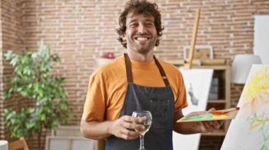 Resimler ve tuvallerle çevrili yaratıcı bir sanat stüdyosunda şarap içen neşeli bir adam.