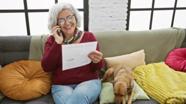 Gri saçlı kadın telefonda konuşuyor, renkli oturma odasındaki kanepede yanında köpeğiyle evrak tutuyor..