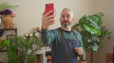 Yakışıklı sakallı adam çiçekçide yeşil bitkilerin arasında selfie çekiyor..