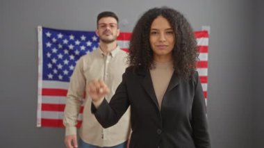 Gülümseyen bir kadın ve arka plandaki bir adam, dayanışma ya da protesto göstergesi olarak yumruklarını kaldırıyor, kapalı bir ofiste Amerikan bayrağını sergiliyor..