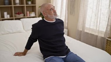 Sakallı, sıradan giyinmiş olgun kel bir adam, iyi aydınlatılmış bir yatak odasındaki beyaz yatakta düşünceli bir şekilde oturur.