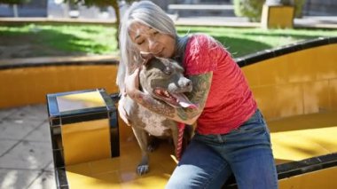 Neşeli orta yaşlı bir kadın, yeşil saçlı köpeğine neşeyle sarılırken, güvenli bir şekilde bir bankta oturuyor, yeşil bir açık hava bahçesinin kalbinde saf mutluluk yayıyor..