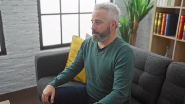 Sakallı, rahat ve olgun bir adam modern bir oturma odasında rahatça oturuyor, rahatlığını ve rahatlığını ifade ediyor..