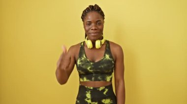 Neşeli Afro-Amerikalı kadın spor giyim ve kulaklık takıyor, sarı arka planda izole edilmiş, ışıl ışıl bir gülümsemeyle kendinden emin bir onay işareti sergiliyor.