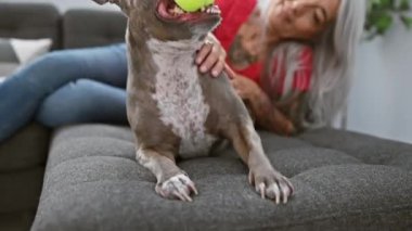 Orta yaşlı, kır saçlı, köpeği sırıtan, kendine güvenen, evinde oynayan bir kadın.