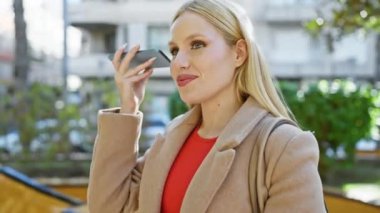 Şehir parkında yeşillik içinde akıllı telefon kullanan sıradan sarışın kadın.