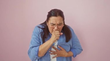 Sağlıksız orta yaşlı, gözlüklü, pembe arka planda öksüren İspanyol bir kadın. Soğuk algınlığı veya bronşit belirtileri.