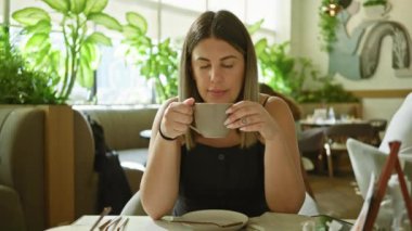 Güneşli, bitkilerle dolu bir kafede kahve içen güzel genç bir kadın.