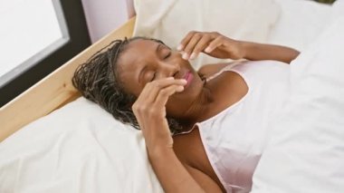 Afro-Amerikalı bir kadın yatağında uzanıyor, yüzüne dokunuyor, yatak odası ortamında rahatsızlık ya da acı veriyor..