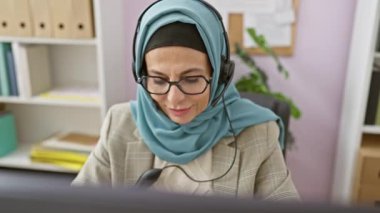 Başörtüsü ve kulaklık takan neşeli bir kadın modern ofis ortamında çalışırken gülümsüyor..