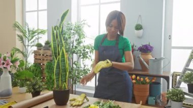 Önlüklü Afrikalı Amerikalı bir çiçekçi, çiçekçi ve saksılarla çevrili bir çiçekçide eldivenlerini çıkarıyor..