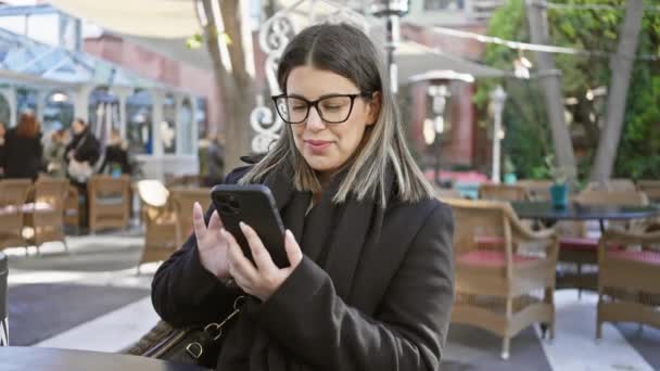 Μια Χαμογελαστή Νεαρή Γυναίκα Γυαλιά Χρησιμοποιώντας Ένα Smartphone Ενώ Κάθεται — Αρχείο Βίντεο