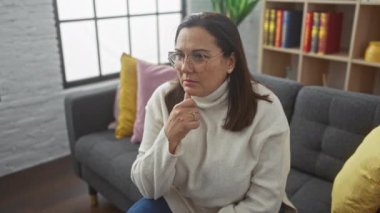 Düşünceli orta yaşlı bir kadın modern bir oturma odasında oturmuş düşünceli bir ifadeyle.