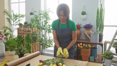 Afro-Amerikalı bir kadın çiçekçide çiçek düzenliyor. Etrafı bitki ve çömleklerle çevrili..