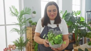 Güneşli bir çiçekçinin içinde saksı saksısı tutan gülümseyen bir kadın. Çeşitli yeşilliklerle süslenmiş.