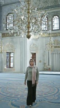 Gözlüklü, tesettürlü, tesettürlü bir kadın süslü bir İstanbul camisinin içinde karmaşık İslami dizaynlar ve avizelerle duruyor..