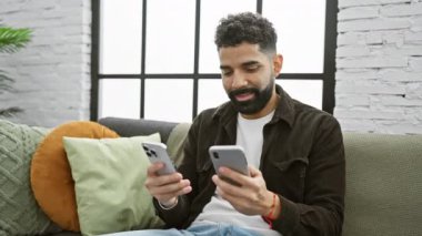 Sakallı genç bir adam, rahat bir oturma odasında iki akıllı telefonuyla birden fazla iş yapıyor, rahat ama nişanlı bir tavır sergiliyor..