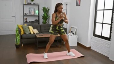 Modern bir oturma odasındaki pembe yoga minderinin üzerinde egzersiz yapan Afrikalı Amerikalı kadın..
