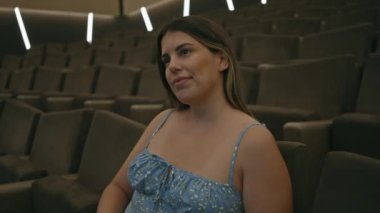 Gülümseyen genç bir kadın geniş bir sinema salonunda lüks koltuklar ve modern ışıklandırmayla eğleniyor..