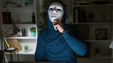 Maske takan ve kapüşonlu genç bir adam modern bir ofiste dizüstü bilgisayarla oturuyor, gizem ve anonimliği çağrıştırıyor..