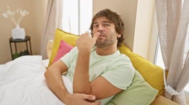 Gururlu, beyaz bir adam, pijama giyiyor, kameraya gülümsüyor, kollarını kavuşturup, elini çenesine koyuyor, düşünceli, pozitif bir ruh hali içinde, rahat yatak odasında..