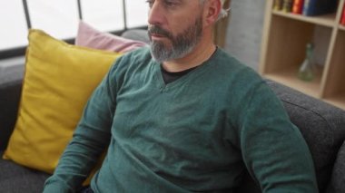 Kafkasyalı orta yaşlı sakallı bir adam, evinde gri bir kanepede otururken göğsünü tutuyor..