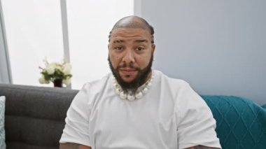 Neşeli Afrikalı Amerikalı adam gülümsüyor ve el pençe divan oturuyor, parmaklarının arasından eve bakıyor..